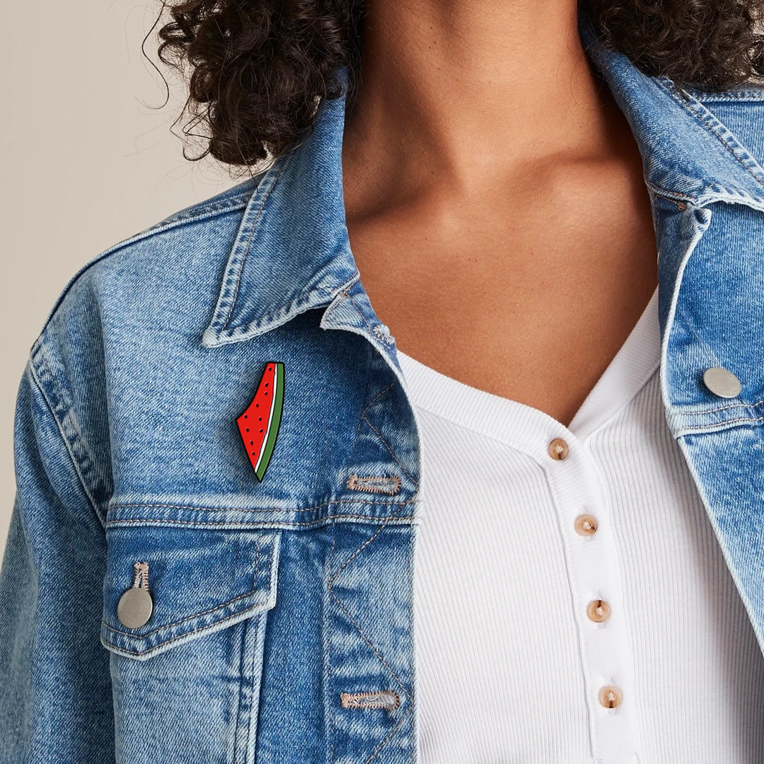 Palestine Watermelon Enamel Label Pin on a blue jean jacket worn by a female model with curly black hair. Sold by the Classy Vendor.